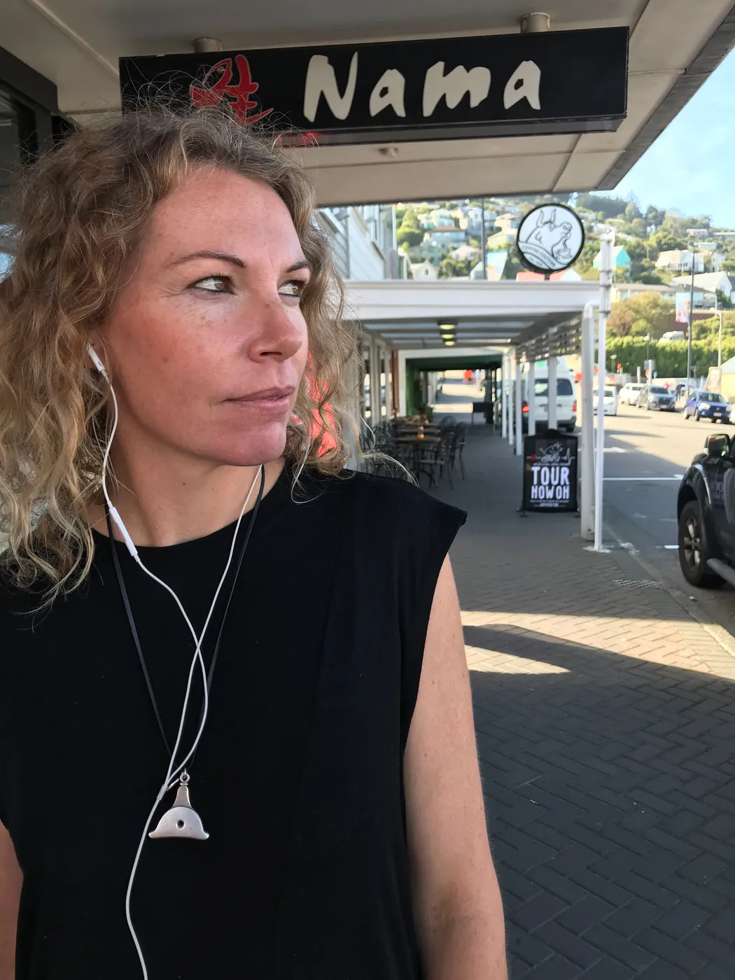 A woman wearing earphones stands on a shopfront street, listening