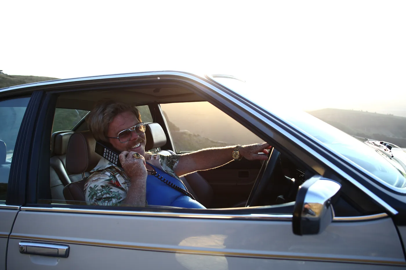 A man in sunglasses and a floral shirt talks on a car phone from the driver's seat of an old sedan