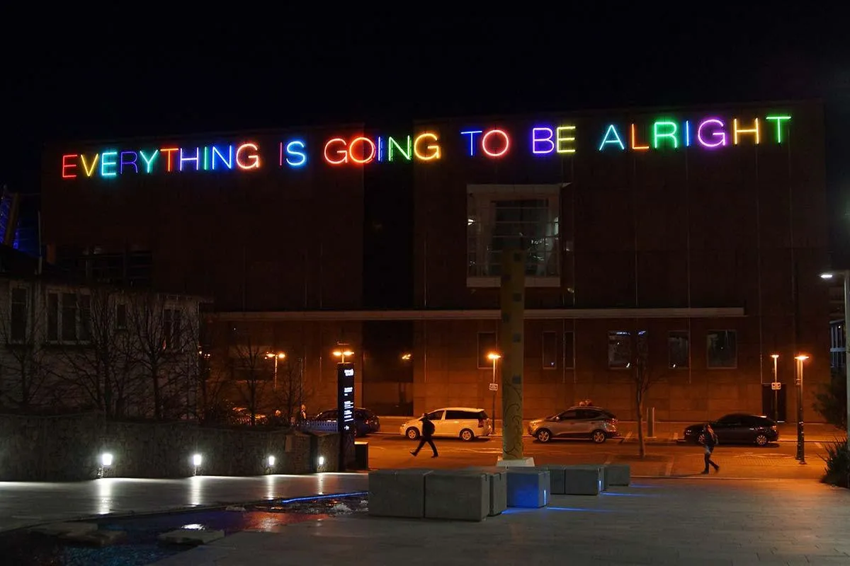 Neon letters spelling Everything is going to be alright glow across a building at night
